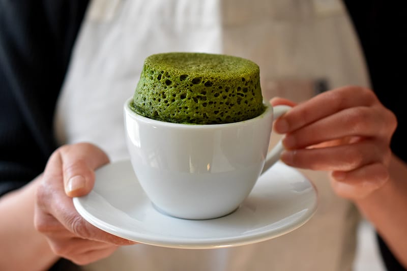 A woman is holding a white coffee mug and saucer. The mug is filled a puffed up green matcha mug cake. 