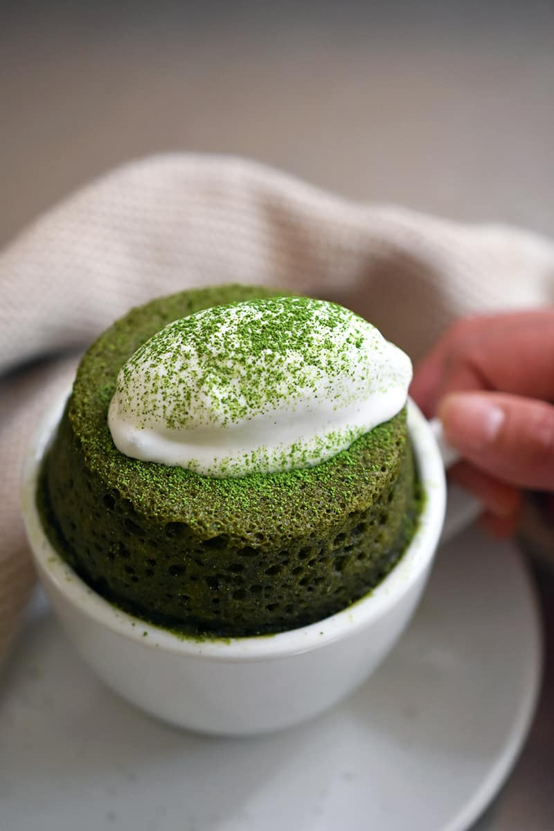 A hand holding the handle of a white mug filled with paleo matcha mug cake and topped with dairy-free whipped coconut cream. 