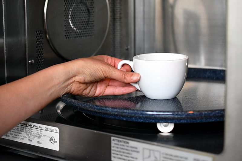 A hand placing a white coffee mug filled with paleo matcha mug cake into an open microwave.